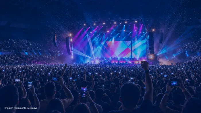 Arena de shows lotada com luzes de palco coloridas e telão ao fundo, imagem meramente ilustrativa