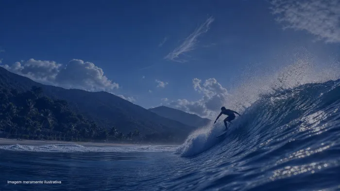 Surfista em silhueta dropando onda em praia de mar aberto com morros ao fundo, imagem meramente ilustrativa