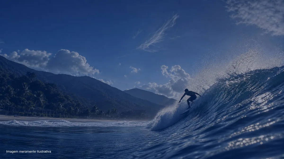 Surfista em silhueta dropando onda em praia de mar aberto com morros ao fundo, imagem meramente ilustrativa