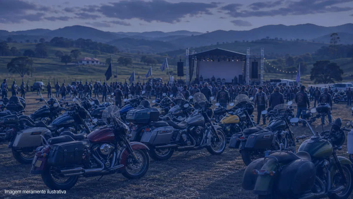 Dezenas de motocicletas estacionadas em um campo, com palco montado ao fundo, imagem meramente ilustrativa